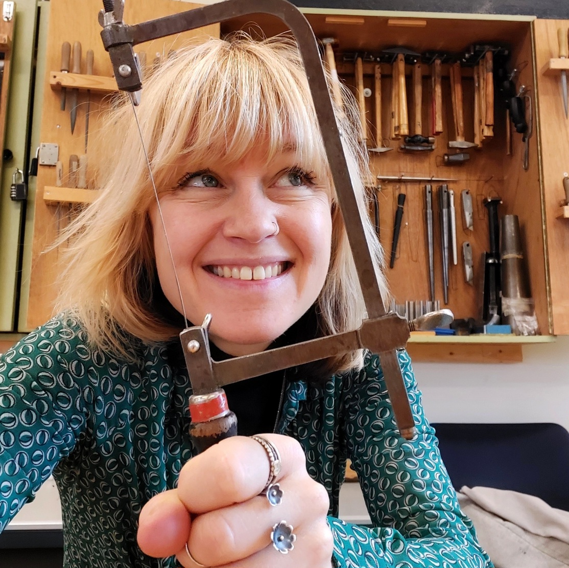 Docent Sacha Ofman houdt lachend een figuurzaag omhoog in haar edelsmeedatelier vol gereedschap