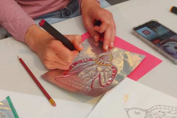 Deelnemer werkt aan een geluksamulet door een vogelmotief in metaalfolie te drijven en in te kleuren tijdens de workshop