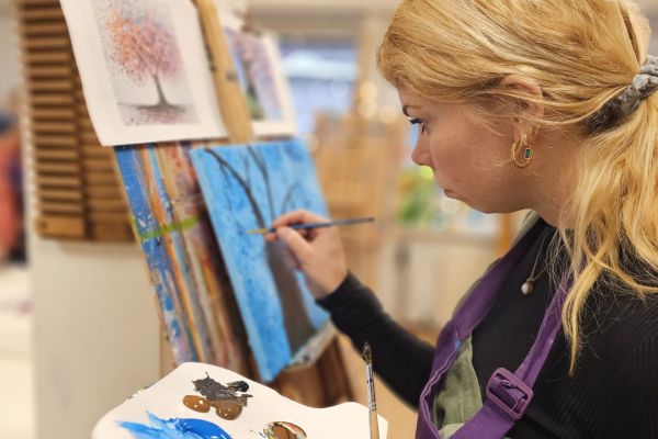 Cursist schildert met acrylverf een boom op doek aan de ezel, met palet in de hand, in het creatief atelier